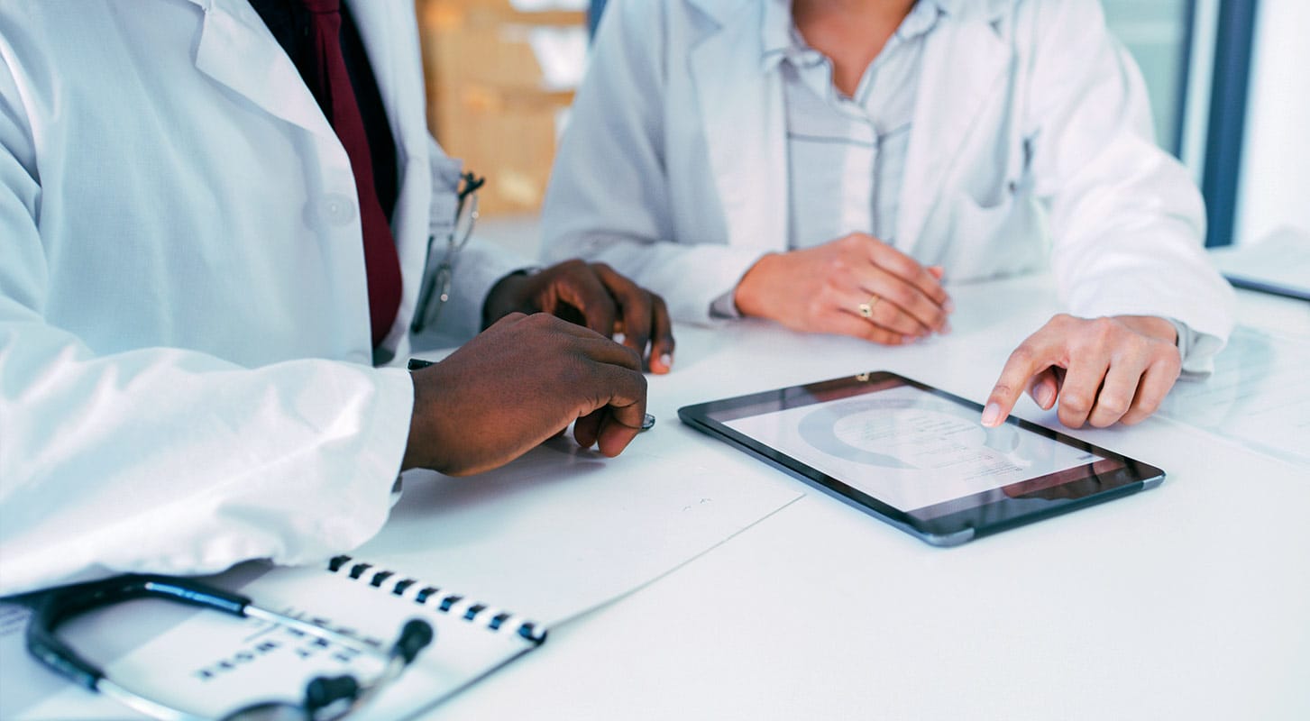 Two doctors in white coats reviewing patient data on a digital tablet at a table, with a stethoscope and notebook in the foreground.